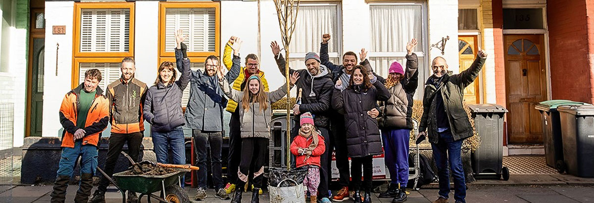 Group of people standing on a pavement in residential area waving at the camera. There is an unplanted tree & wheelbarrow in the centre of the frame